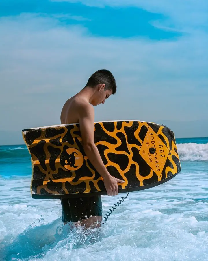 A young man stands in the surf holding a colorful bodyboard under a bright blue sky.