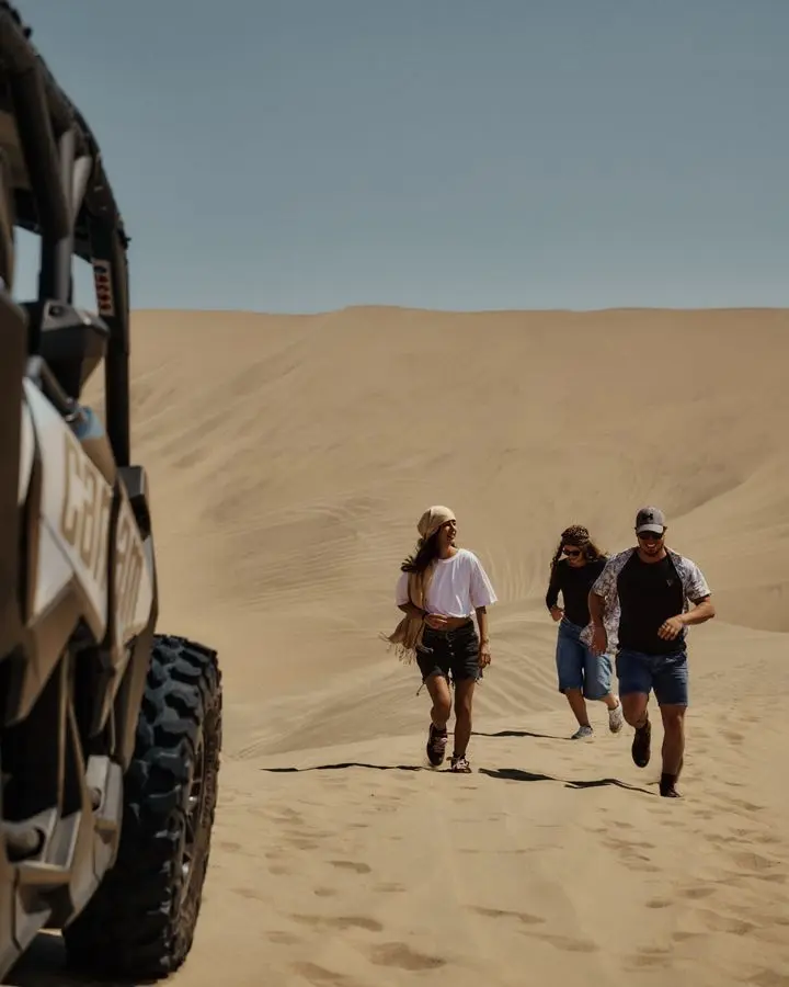 Three friends having fun running in desert dunes near off-road vehicle.