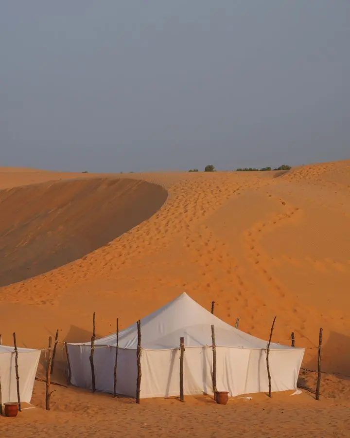 White nomadic tents set amidst the golden sand dunes of a vast desert under a clear blue sky.