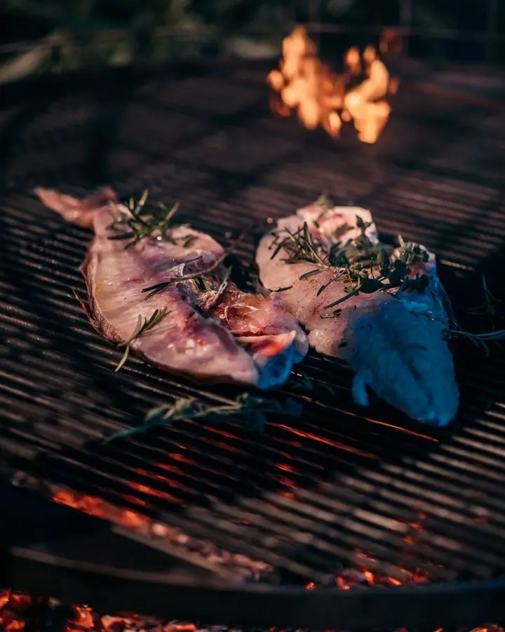 Close-up of fish slices being grilled with herbs over an open flame, highlighting outdoor cooking.