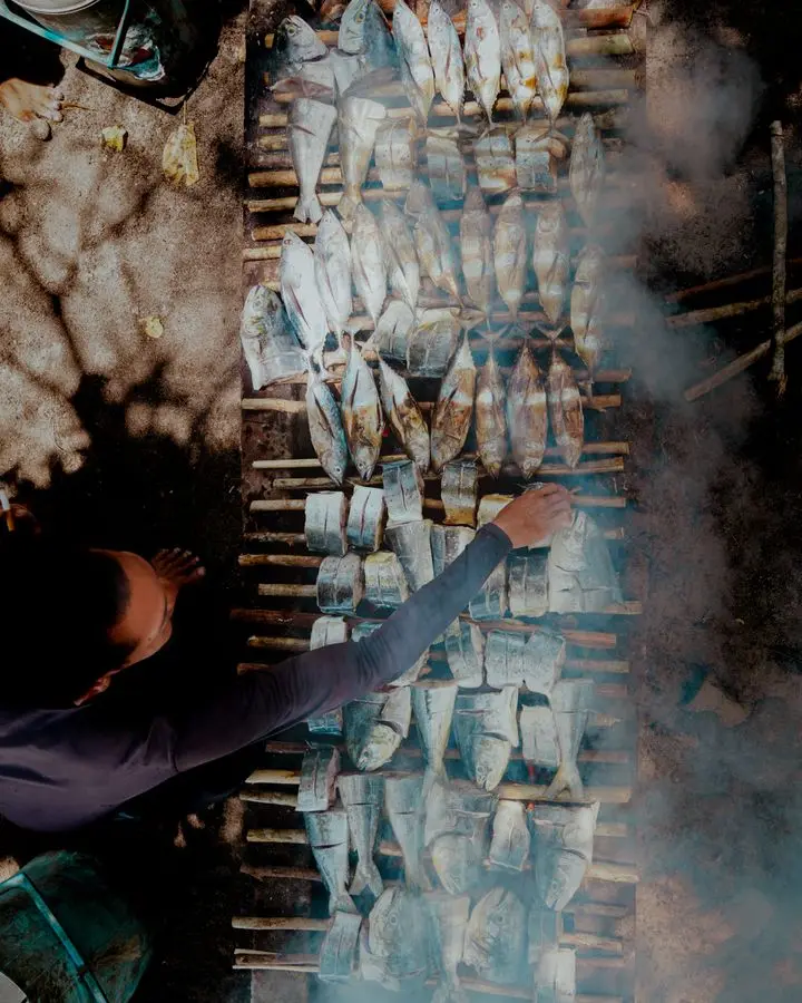 Person preparing fish on an outdoor grill with smoke and shadows.