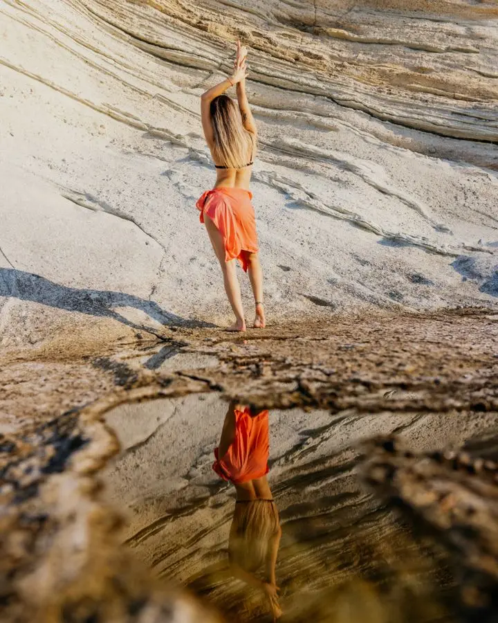 A woman in a red wrap practicing yoga on a rocky beach, reflecting in a tide pool.