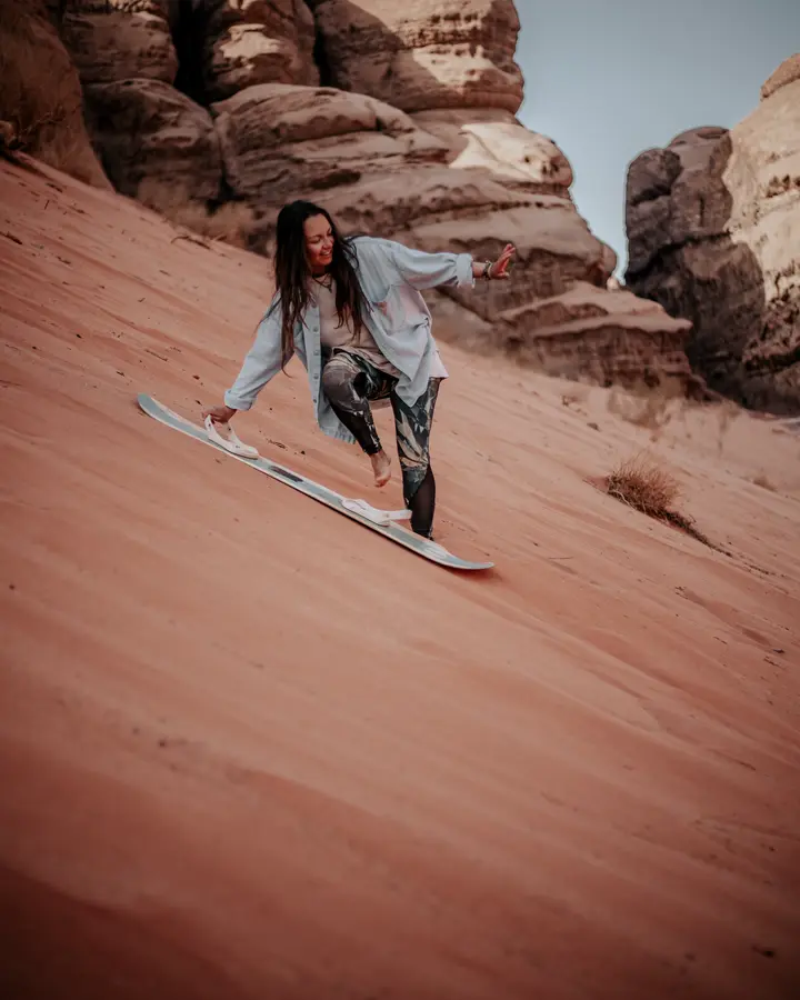 A woman enjoying sandboarding down a steep desert dune with rocky formations in the background.