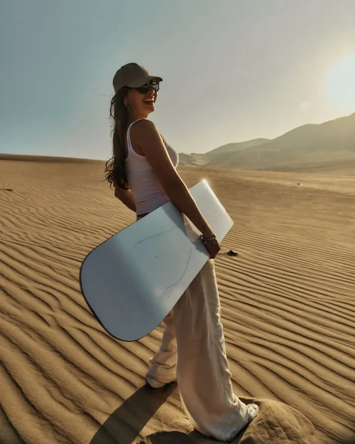 A woman holds a sandboard on a sunny desert dune, ready for adventure.